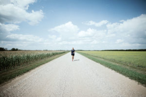 Karen Edwards runs along a wide road of prairie, with blue skies in the background and green grass on either side.