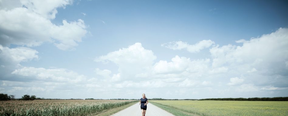 Karen Edwards runs along a wide road of prairie, with blue skies in the background and green grass on either side.