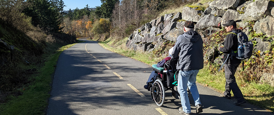 Two adults walking on a paved trail. One adults is guiding another in a wheelchair. 
