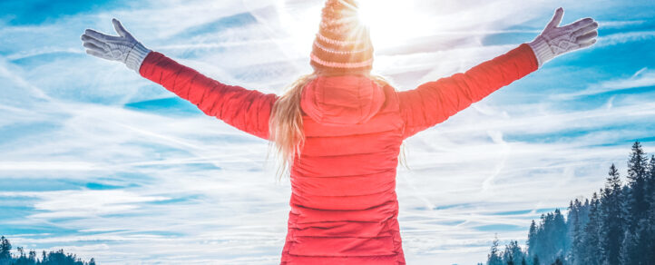 A woman in an orange down-filled jacket, gloves, and a toque, back the the camera, facing the sunshine on a beautiful day.