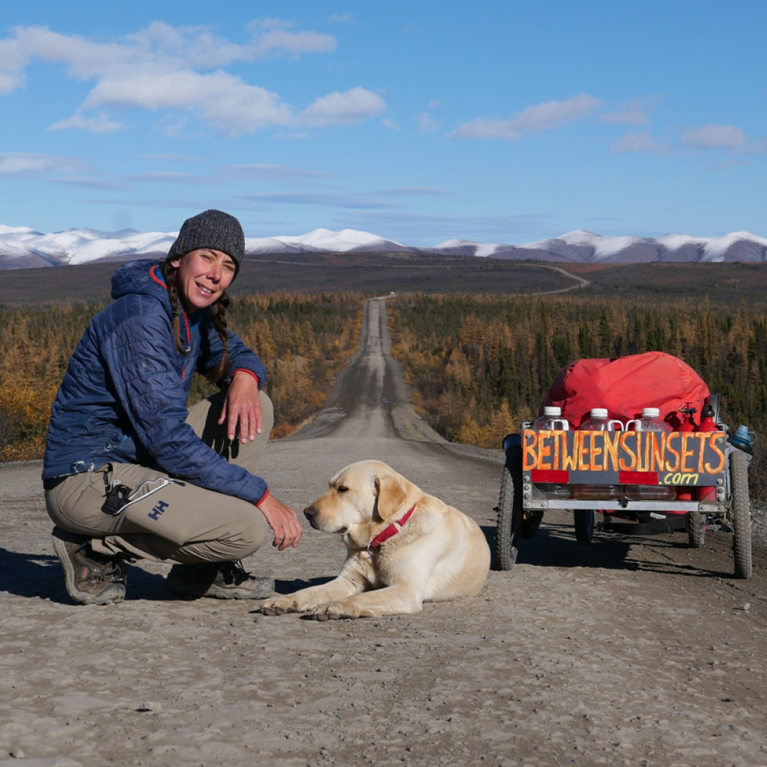 Melanie Vogel with her dog, kneeling on a path in a remote area, with mountains and forest in the background. There's a wagon with her, and sign on the back of it says between sunsets dot com. Melanie Vogel avec son chien, à genoux sur un chemin dans une région reculée, avec des montagnes et une forêt en arrière-plan. Elle est accompagnée d'un chariot, dont le panneau à l'arrière indique "between sunsets dot com".