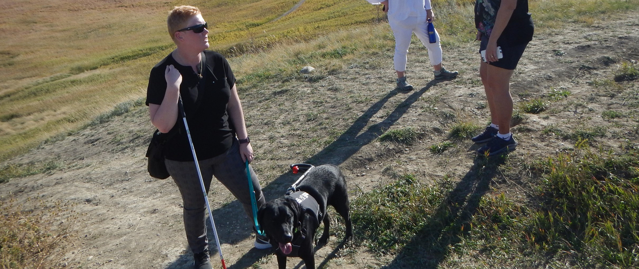 An adult on the grass holding a leash with a dog in one hand and a guide can in the other. 