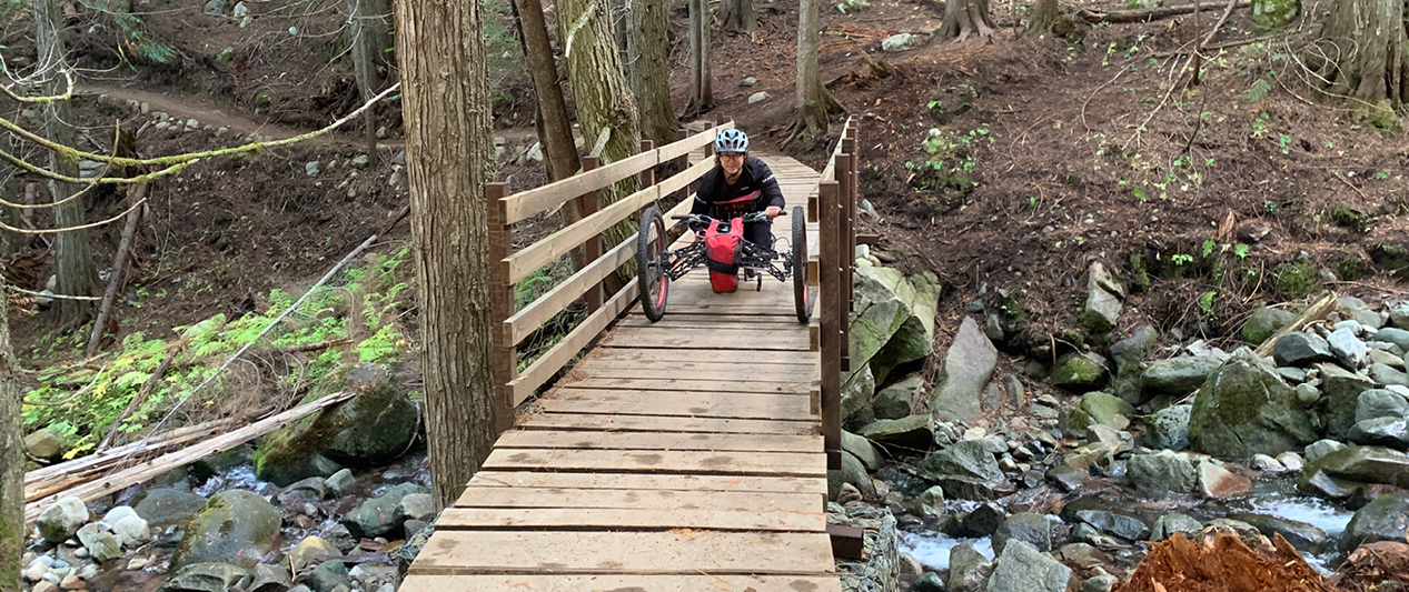 An adult on an adaptive mountain bike at the Morning Mountain Recreation Area, BC, trail on a wooden bridge. 