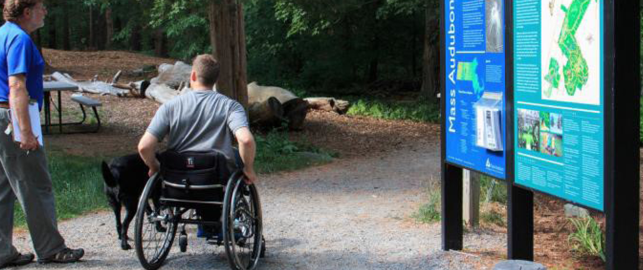 The back of an adult in a wheelchair on the trail with a dog beside him. An adult beside them is holding a clip board