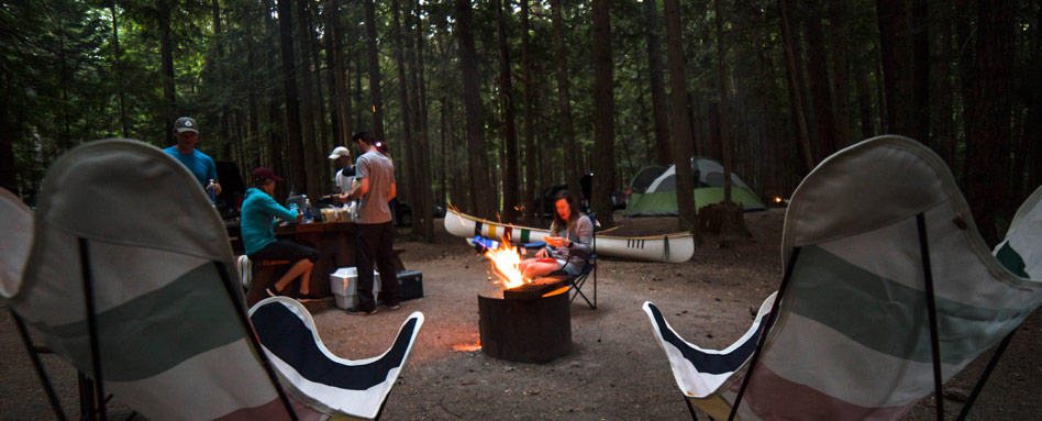 A campfire setup with a group of people, outdoor chairs and picnic table surrounding the fire pit. This is an example of Leave No Trace Principles: Minimize campfire impacts.