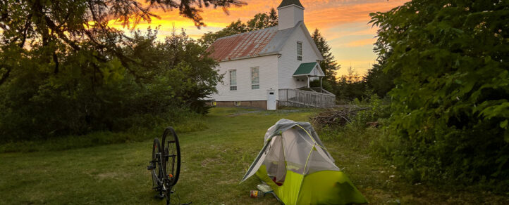 A tent and a bike on the grass in front of an older house with a sunset in the background