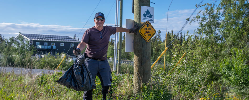 Volunteer at the Klondike Trail Care Event holding a garbage bag and standing by the Trans Canada Trail sign