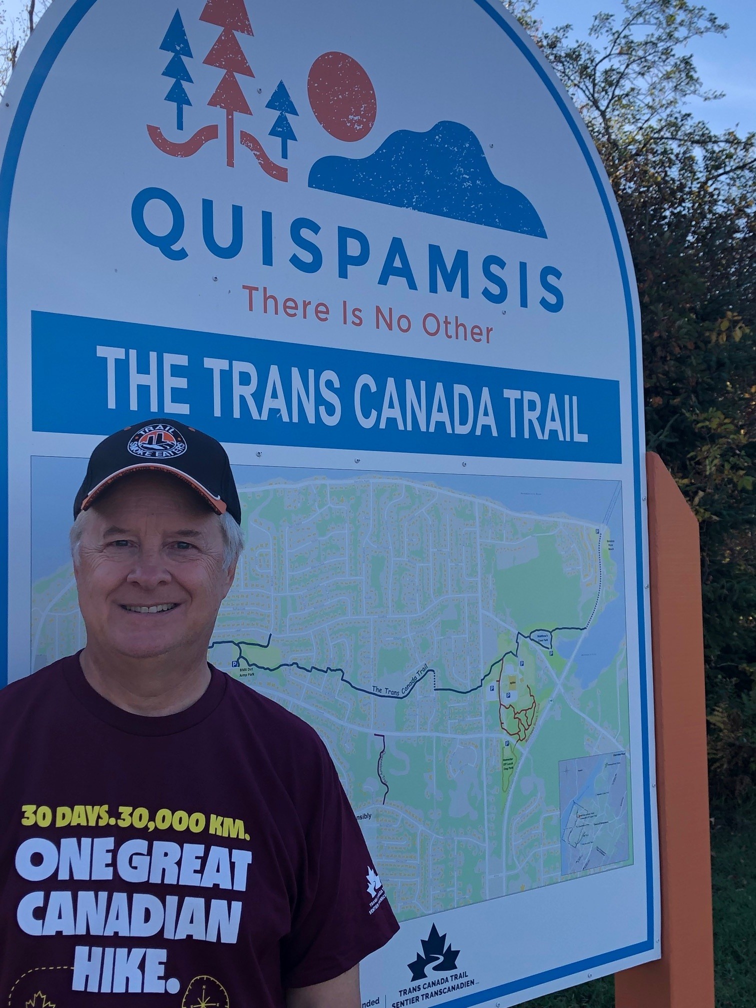 Person in a burgundy shirt and cap stands in front of a large trail sign for The Trans Canada Trail in Quispamsis, with a map displayed behind them.