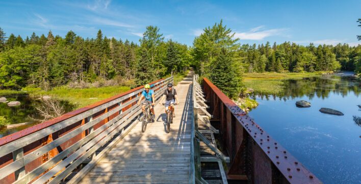 Two people cycle across a wooden bridge spanning a small body of water.