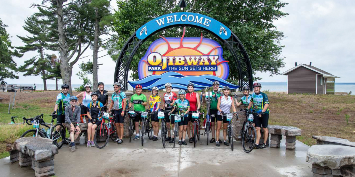 Cyclists of the Great Waterfront Trail Adventure standing in front of the welcome sign for Ojibway Park sign