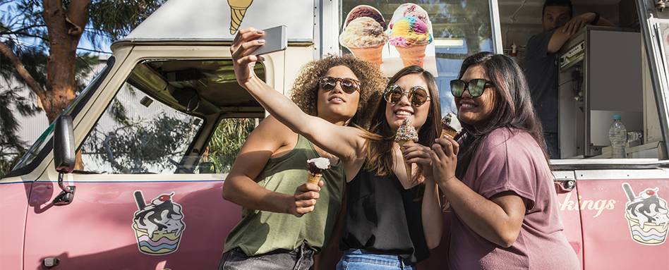 Three women smiling and taking a selfie in front of an ice cream truck. Trois femmes souriantes prennent un selfie devant un camion de glaces. 