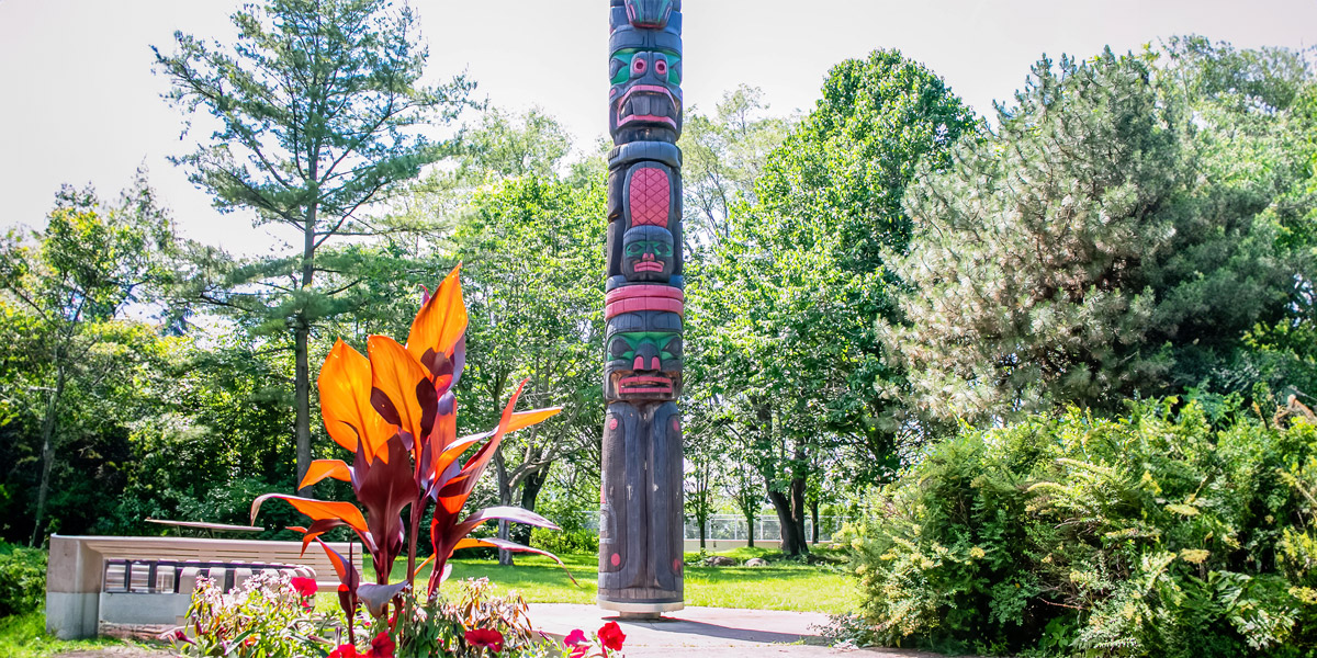 A totem pole stands tall in a park, surrounded by vibrant flowers, showcasing cultural artistry amidst nature's beauty. Un totem se dresse fièrement dans un parc, entouré de fleurs éclatantes, mettant en valeur l'art culturel au milieu de la beauté de la nature.