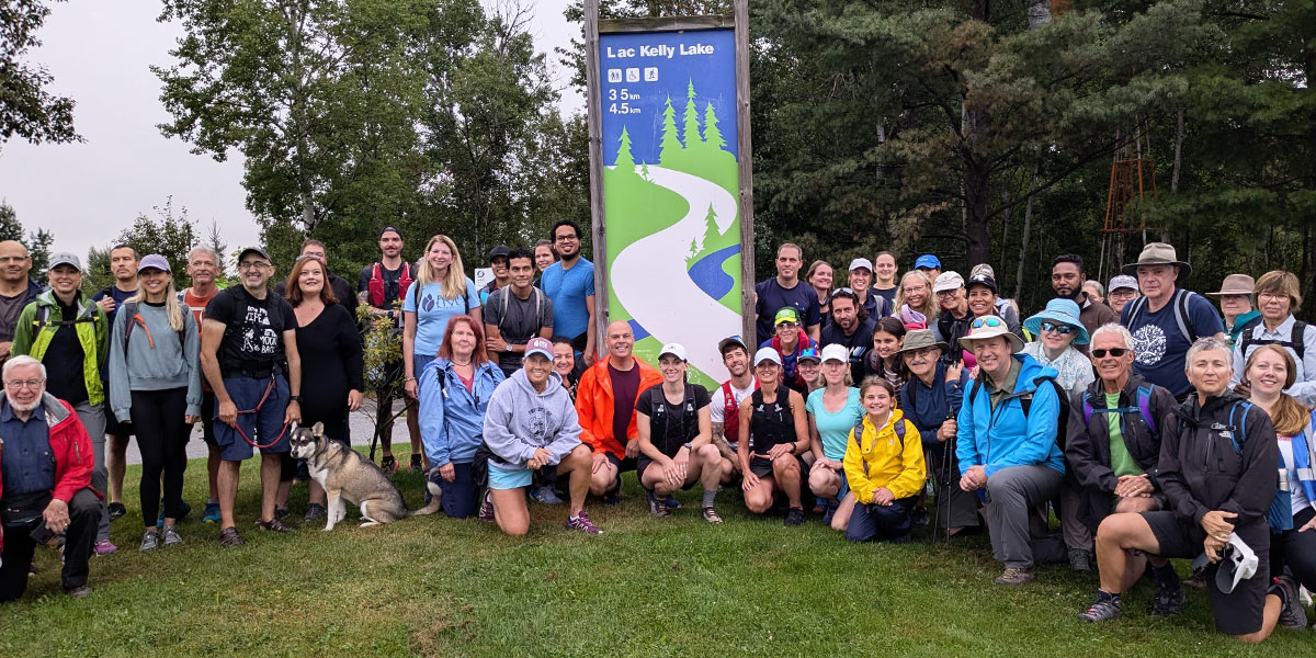 A diverse group of individuals smiling and posing together in front of a large sign, capturing a moment of camaraderie. Un groupe diversifié d'individus souriant et posant ensemble devant un grand panneau, capturant un moment de camaraderie.