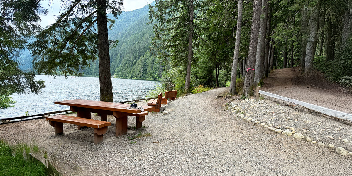 A picnic table and bench on a lakeside trail, designed for accessibility and enjoying nature's beauty. Une table de pique-nique et un banc sur un sentier au bord du lac, conçus pour être accessibles et profiter de la beauté de la nature.