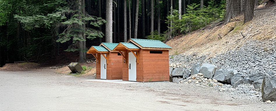 Two wooden outhouses located beside a dirt trail, set against a backdrop of greenery. Deux dépendances en bois situées à côté d'un chemin de terre, sur fond de verdure.