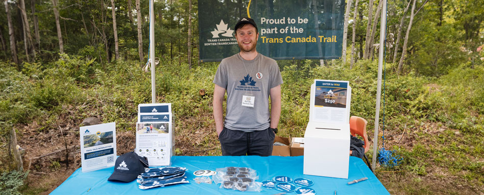 A Trans Canada Trail staff member stands behind a table with a sign reading "Proud to be part of the Trans Canada Trail." Un membre du personnel du sentier Transcanadien se tient derrière une table sur laquelle est affiché un panneau indiquant « Proud to be part of the Trans Canada Trail »