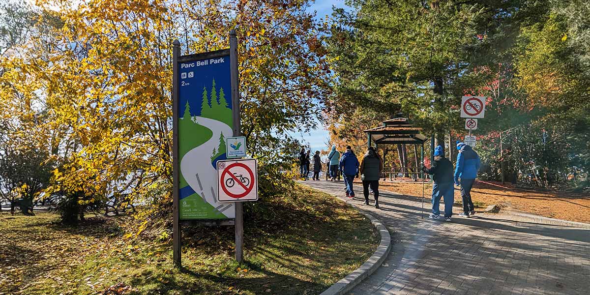 People walking along a path near a "Parc Bell Park" sign, surrounded by greenery and a clear sky. Des personnes marchent le long d’un sentier près d’un panneau indiquant « Parc Bell Park », entourées de verdure sous un ciel dégagé.