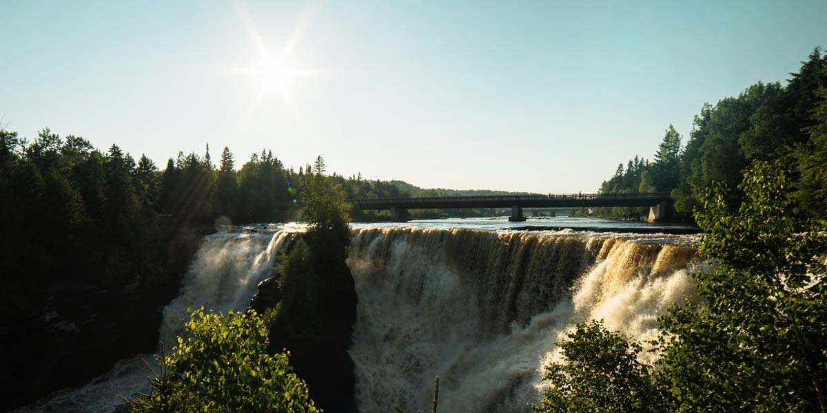 A bright sun illuminates a serene waterfall cascading through a lush forest, creating a picturesque natural scene. Un soleil éclatant illumine une cascade sereine qui coule à travers une forêt luxuriante, créant une scène naturelle pittoresque.