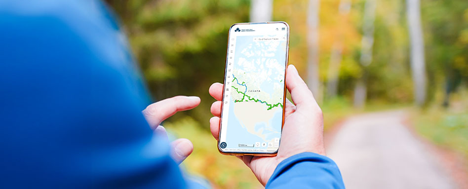 A person displays a smartphone featuring the Trans Canada Trail map on its screen, highlighting outdoor navigation. Une personne montre un téléphone intelligent sur lequel est affichée la carte du Sentier transcanadien, mettant en valeur la navigation en plein air.