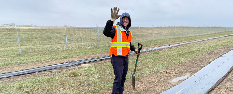 A man in an orange vest and safety attire stands on a field, surveying the area around him. Un homme portant un gilet orange et une tenue de sécurité se tient debout sur un terrain, observant la zone qui l'entoure.