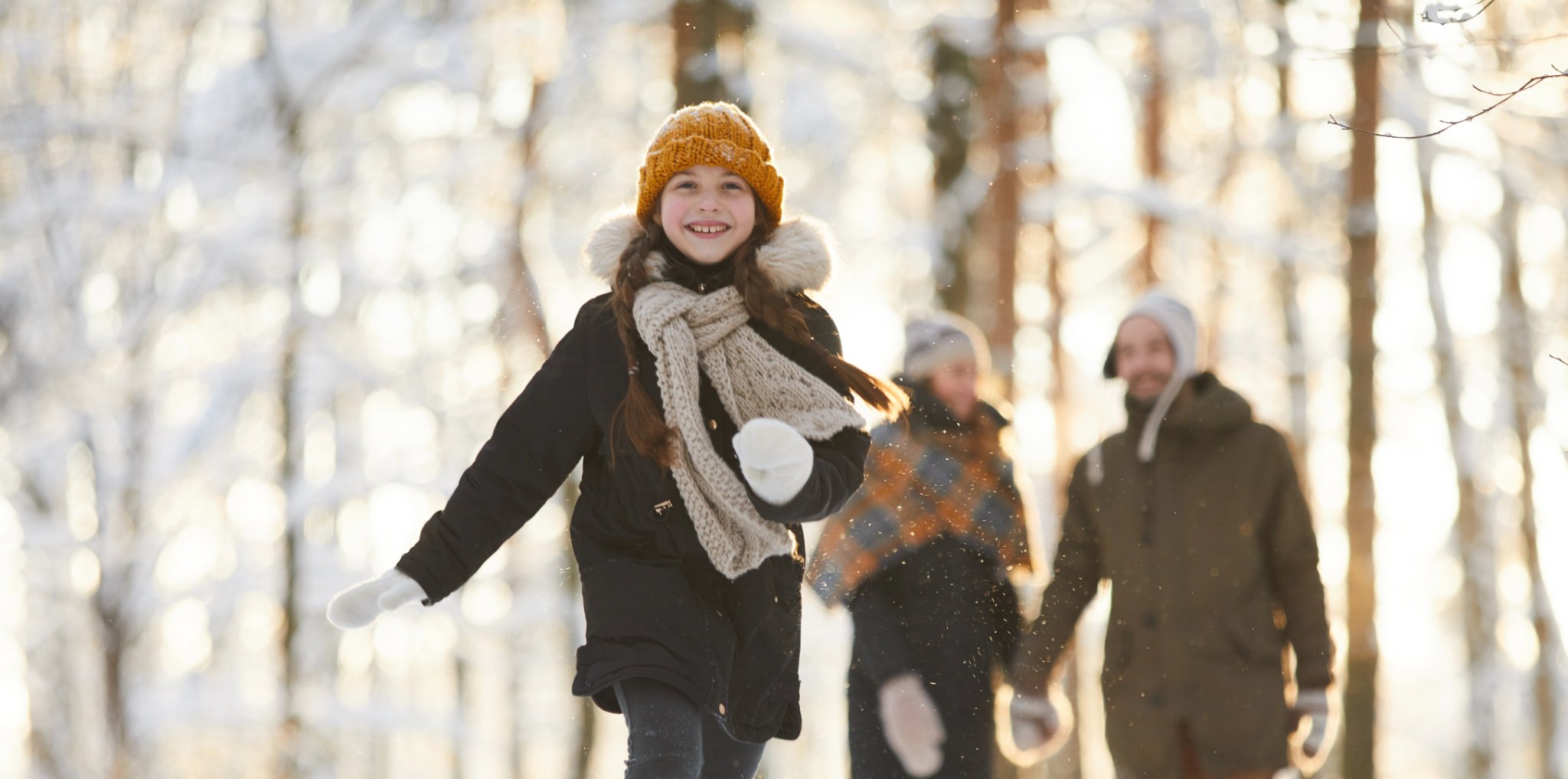 Winter time in the forest. A beautiful sunny day. A little girl wearing a toque and scarf runs towards the camera. Her parents are in the background.