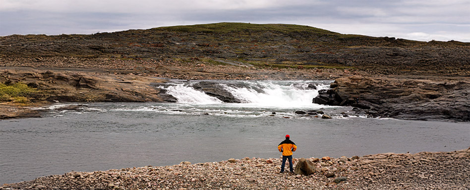 A person stands on a rock along the Itijjagiaq Trail, surrounded by natural beauty and scenic views. Une personne se tient debout sur un rocher le long du sentier Itijjagiaq, entourée de beautés naturelles et de vues panoramiques.