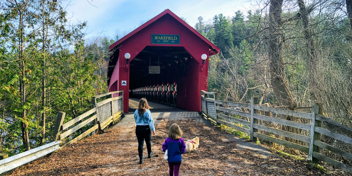 Two children stroll across the covered bridge on Wakefield Trail, surrounded by nature and sunlight. Deux enfants se promènent sur le pont couvert du tronçon de Wakefield, entourés par la nature et baignant dans la lumière du soleil.