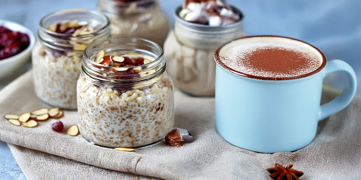 A mason jar filled with cranberry-almond overnight oats, topped with cinnamon and nuts, alongside spiced hot chocolate mix. Un pot Mason rempli de flocons d'avoine aux canneberges et aux amandes préparés la veille, garni de cannelle et de noix, accompagné d’un mélange pour chocolat chaud épicé.