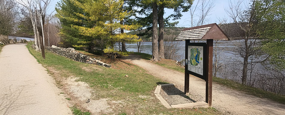 A signpost indicating a scenic trail along a river, inviting visitors to enjoy a storybook walk in nature. Un poteau indicateur signalant un sentier pittoresque longeant une rivière, invitant les visiteurs à une promenade bucolique en pleine nature.