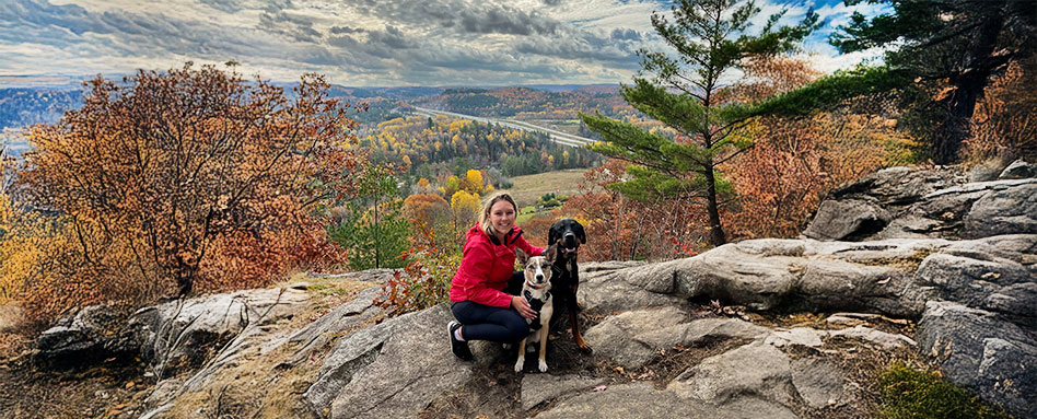 A woman perched on a rock with her two dogs, enjoying a serene moment in a natural setting, with greenery in the background. Une femme assise sur un rocher avec ses deux chiens, savourant un moment paisible dans un cadre naturel, avec de la verdure en arrière-plan.