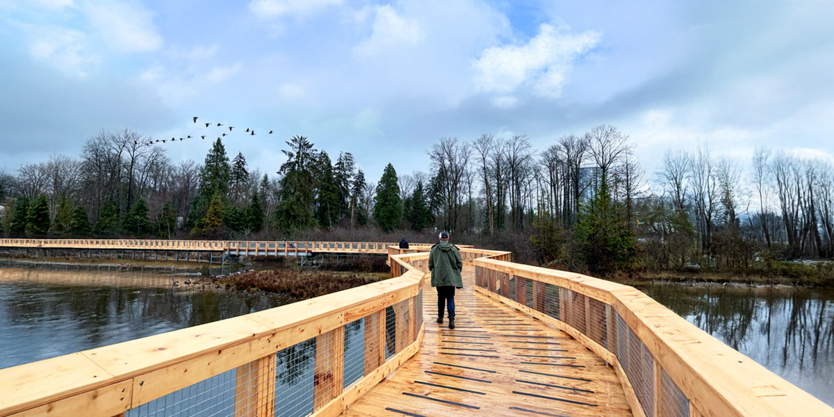 Individual walking on a wooden bridge, enjoying the tranquil lake views along a scenic trail in Vancouver. Une personne marche sur un pont en bois, profitant de la vue paisible sur le lac le long d’un sentier panoramique à Vancouver.