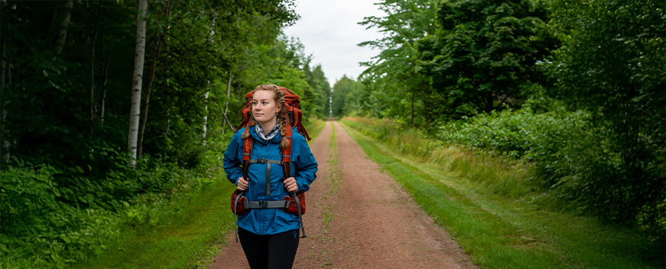 A woman with a backpack strolls along a dirt road on Prince Edward Island, surrounded by natural beauty. Une personne portant un sac à dos se promène sur un chemin de terre sur l’Île-du-Prince-Édouard, entourée de la beauté naturelle des lieux.