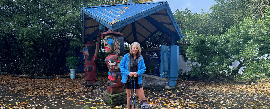 Mary Charleson stands in front of a blue gazebo and a totem pole, showcasing a serene outdoor setting. Mary Charleson se tient devant un pavillon bleu et un totem, mettant en avant un environnement extérieur serein.