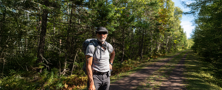 A man carrying a backpack stands on a dirt road in Prince Edward Island, with scenic views of nature in the background. Une personne portant un sac à dos se tient sur un chemin de terre sur l’Île-du-Prince-Édouard, avec un panorama naturel à couper le souffle en arrière-plan.