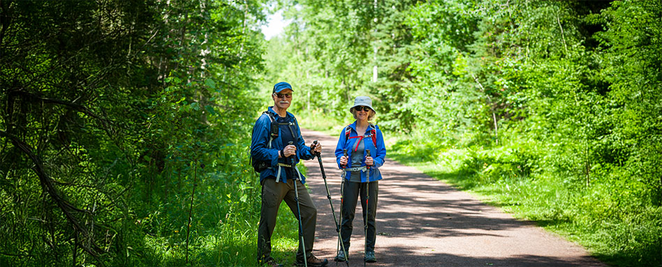 Two individuals with backpacks and walking sticks are walking along a dirt road on an island surrounded by natural scenery. Deux personnes munies de sacs à dos et de bâtons de marche avancent le long d’un chemin de terre sur une île, immergées dans un décor naturel enchanteur.