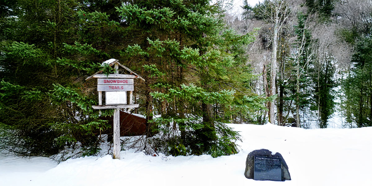 A sign in the snow near a tree, marking the snowshoe trail in Sault Ste. Marie, surrounded by a winter landscape. Un panneau dans la neige près d’un arbre, indiquant le sentier de raquette à Sault Ste. Marie, entouré d’un paysage hivernal.