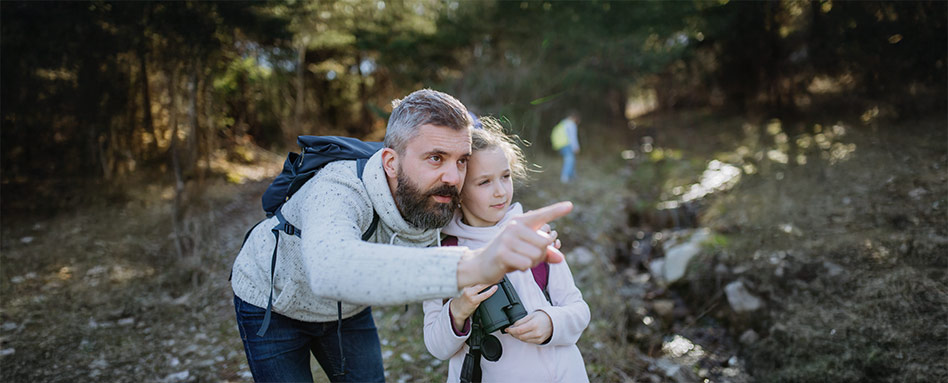 A father and daughter hike through the woods, engaging in birding. Un père et sa fille parcourent un sentier en forêt tout en observant les oiseaux.