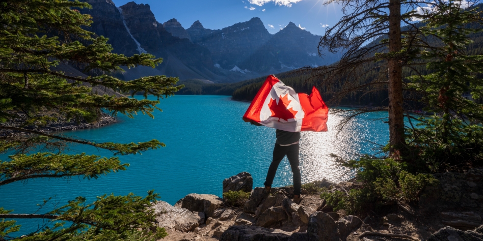 A person standing on a cliff above a lake holding a Canadian flag with mountains in the distance.