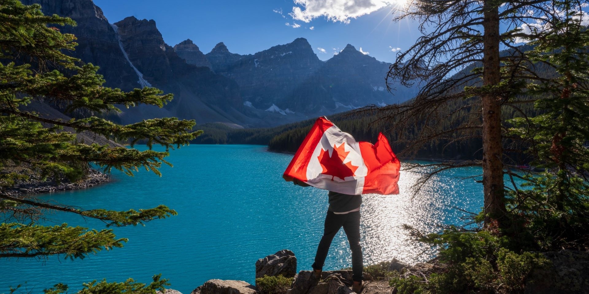 A person holds a Canadian flag on a cliff above a lake with mountains in the background.