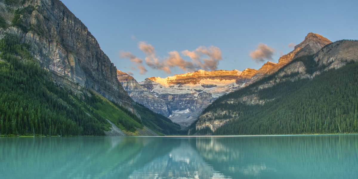 turquoise lake in Banff National Park perfectly mirrors steep, pine?covered mountains and sunrise?lit, snow?capped peaks under a clear blue sky.