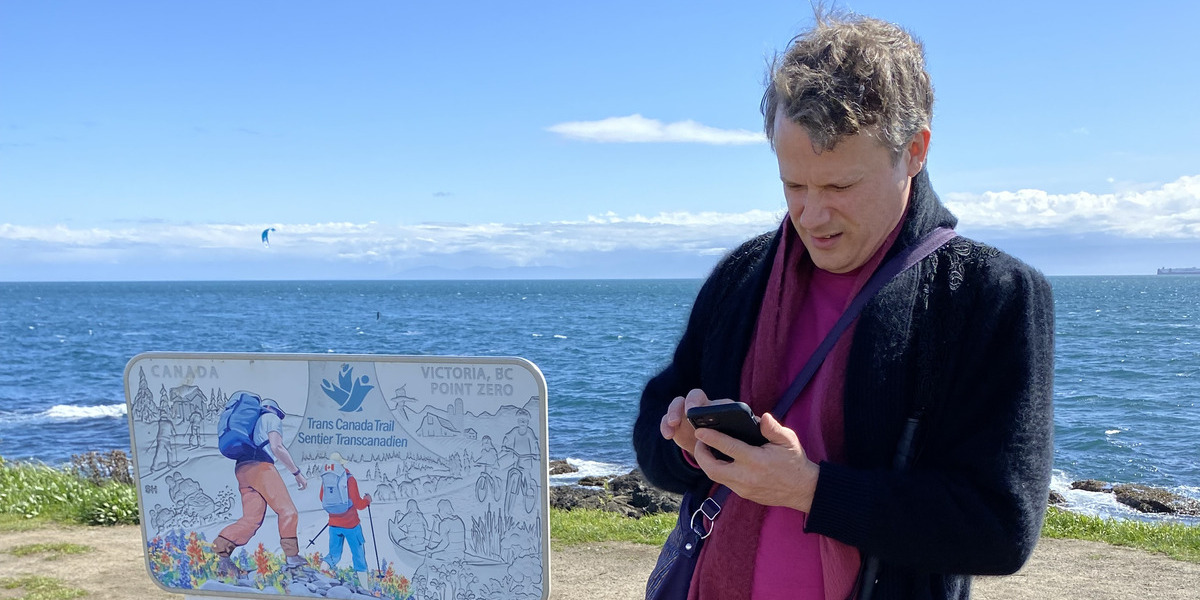 man looking at a phone with trail signage and sea and blue sky behind