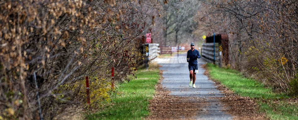 A man wearing sunglasses and a toque running along a trail in late fall. Leaves are on the ground and no branches are on the trees.