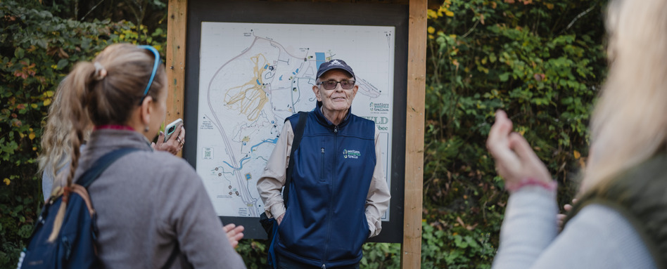 A man stands in front of a trailhead map, looking at two people who are facing him.