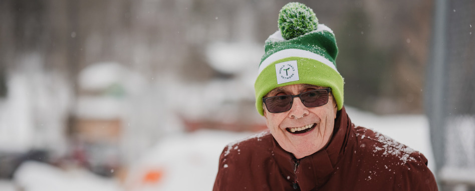2025-02-06_QC_Sentiers-Wakefield-Trails_TCT-B2A-Event | Trans Canada Trail A smiling older man looks out of frame right. He's wearing a green toque and sunglasses. There is snow falling and snow collected on his toque and shoulders.