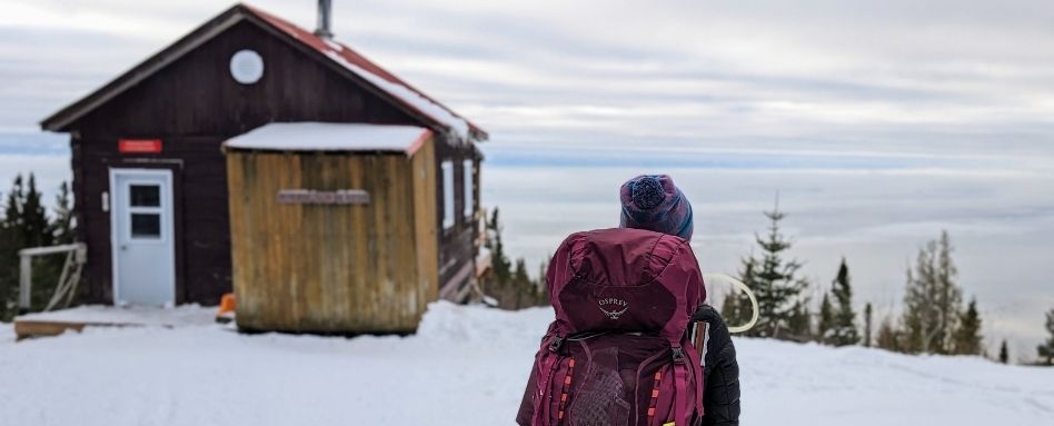 01Feature image_QC_Web story_Famille en Charlevoix_credit Naomie Veillette - ActivMom | Trans Canada Trail A person with their back to the camera on a snowy hill, next to a wood cabin.