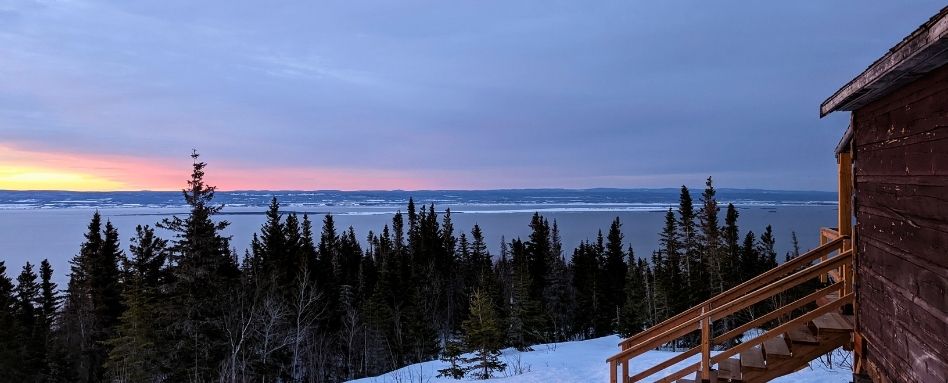 04Support image_QC_Web story_Famille en Charlevoix_credit Naomie Veillette - ActivMom | Trans Canada Trail The sun going down over the treeline and a wintery river. To the right, a wooden hut is partially in frame.