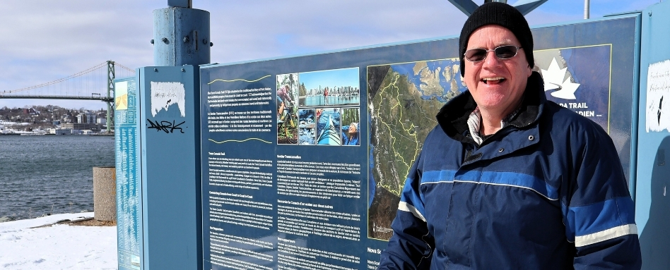 man standing in front of trail signage smiling on a sunny and snowy day