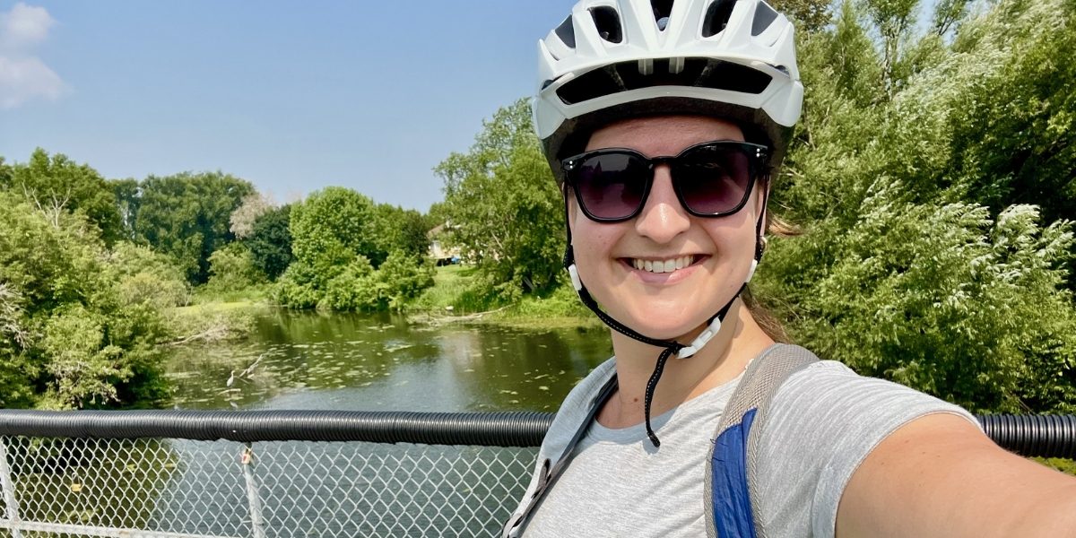 woman taking a selfie in a bike helmet and sunglasses while infront of a lake