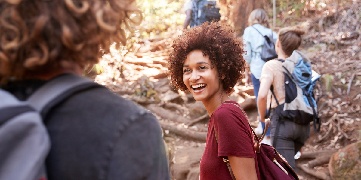 Smiling woman with curly hair in a maroon shirt hiking with a group on a forest trail with backpacks behind her.