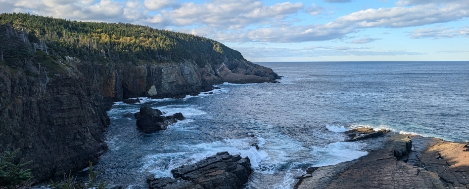 02Support image_Aidan Beckett East_credit Aidan Beckett | Trans Canada Trail Rugged coastal scene with steep, rocky cliffs and evergreen trees, waves breaking on rocks under a blue sky with scattered clouds.